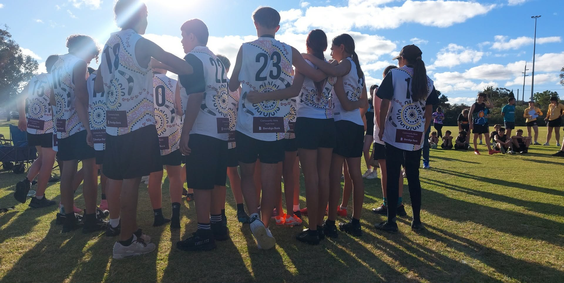 A group of youth in a huddle at Gnoonie Youth Cup with football uniforms on, some with their arms around eachother. Everyone is facing inwards towards the grroup, and away from the camera.